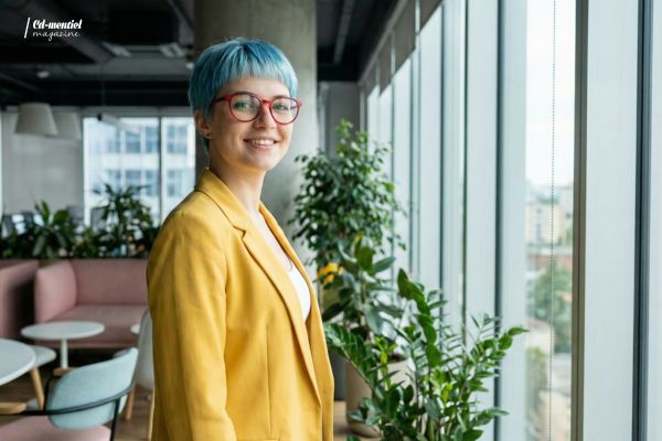 Photo réaliste générée par IA d'une jeune femme entrepreneuse souriante dans un bureau moderne. Elle porte des cheveux bleus courts, des lunettes rouges et un blazer jaune. Image issue du premier test de création de personnage avec Nano Banana Pro.